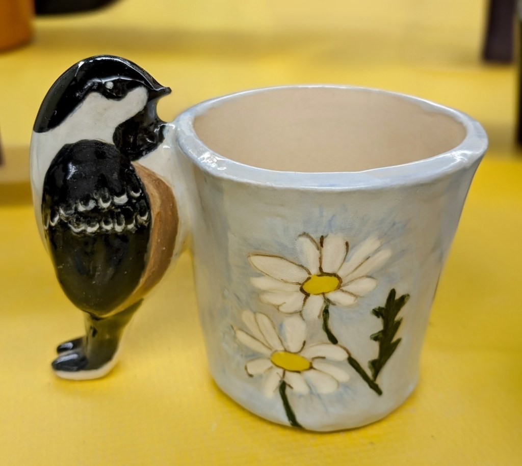 Ceramic chickadee perched beside a pale blue flower pot decorated with daisies.