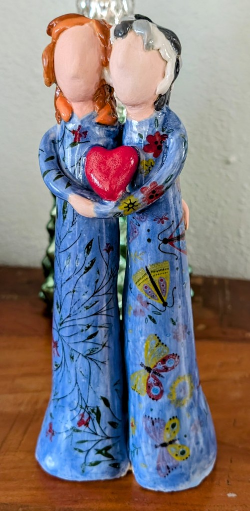 Ceramic figures of Alicia and her mom in blue floral dresses holding a red heart.