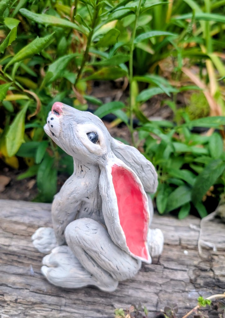 Gray ceramic bunny with pink ears posed outdoors among green plants.